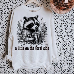 May include: White sweatshirt with a black and white illustration of a raccoon drinking from a cup. The text "a little on the feral side" is printed below the raccoon.