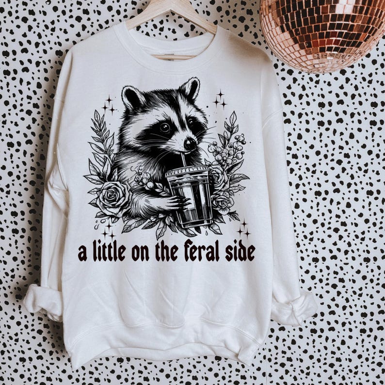 May include: White sweatshirt with a black and white illustration of a raccoon drinking from a cup. The text "a little on the feral side" is printed below the raccoon.