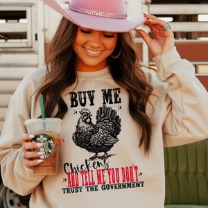 May include: A woman wearing a beige sweatshirt with a black and white chicken graphic and the text "Buy Me Chickens and Tell Me You Don't Trust The Government".