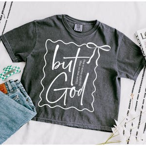 Op de afbeelding: Een donkergrijs T-shirt met een witte afbeelding waarop staat "but God made a way." De afbeelding bevindt zich in een wit kader met een witte strik bovenaan.