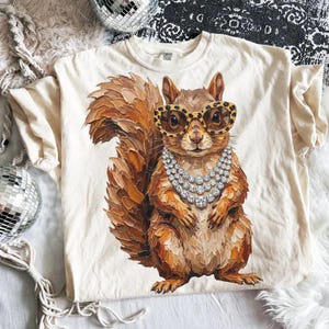 May include: A cream-colored t-shirt featuring a central graphic of a brown squirrel with thick, textured brushstrokes. The squirrel wears large, round leopard-print glasses and a triple-layered necklace made of sparkling, clear, round rhinestones.