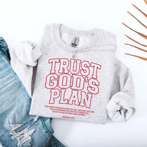 Może przedstawiać: Jasnoszara bluza z napisem "TRUST GOD'S PLAN" w czerwonych literach. Poniżej znajduje się cytat z Jeremiasza 29:11. Bluza jest zestawiona z niebieskimi dżinsami i srebrnym naszyjnikiem.