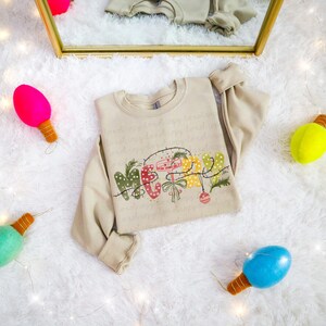 Puede incluir: Una sudadera beige de cuello redondo con la palabra "MERRY" en un diseño festivo y colorido. El diseño incluye elementos verdes, rojos y amarillos, con una guirnalda de luces. La sudadera está doblada sobre una superficie blanca y esponjosa, rodeada de bombillas decorativas.