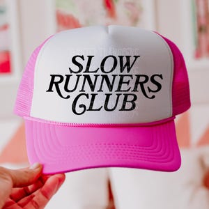 May include: A pink and white trucker hat with the text "SLOW RUNNERS CLUB" in black script. The hat has a pink brim and mesh back, with a white front panel. The hat is a classic style.