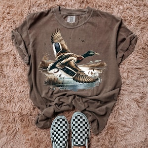 Peut inclure: Un t-shirt marron avec une illustration détaillée d'un canard en vol au-dessus d'un lac. Le canard a des plumes marron, noires et blanches. Le t-shirt est associé à des chaussures à enfiler à carreaux noirs et blancs.