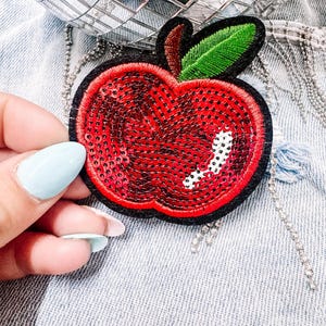 Può includere: Toppa a forma di mela rossa con paillettes, bordo nero e foglia verde. La toppa è su una giacca di jeans. La mela ha un design a pois bianchi. Lo sfondo include una palla da discoteca a specchio e una catena d'argento.