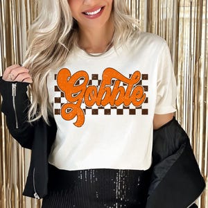 Könnte beinhalten: Weißes T-Shirt mit einem braunen und weißen Karomuster und dem Wort "Gobble" in orangefarbener Schrift.