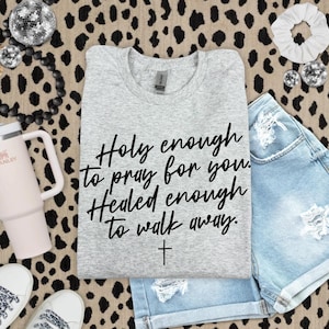 May include: A light grey t-shirt with the text "Holy enough to pray for you. Healed enough to walk away." and a cross graphic. The shirt is styled with denim shorts and accessories on a leopard print background.