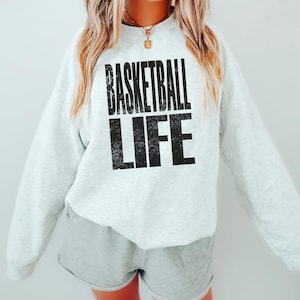 Puede incluir: Una sudadera blanca con el texto "Basketball Life" impreso en negro en la parte delantera. El texto está en una fuente desgastada.