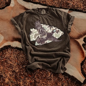 May include: Dark olive green t-shirt featuring a graphic of a duck in flight, set against a camouflage pattern within a stylized shape. The shirt has rolled-up sleeves and a vintage, worn look, suggesting a casual style.