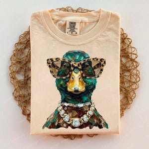 May include: A light peach-colored t-shirt featuring a graphic of a mallard duck wearing leopard-print glasses and a gold chain with square crystals. The shirt has a crew neckline and a Comfort Colors brand label printed on the inside neck area.
