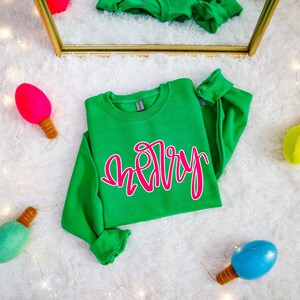 Puede incluir: Una sudadera verde con la palabra "Merry" en escritura rosa. La sudadera se muestra sobre una superficie blanca y esponjosa, con luces decorativas coloridas y un espejo en el fondo. La sudadera tiene mangas largas y cuello redondo.