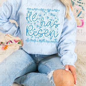 May include: Light blue sweatshirt with the text "He has Risen" in a blue script font. The text is set against a floral pattern with blue flowers and bows. The sweatshirt is a casual style.