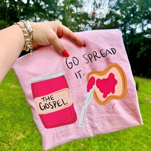 以下が含まれることがあります： 「GO SPREAD IT.」の文字と、ジャムの瓶とジャムを塗ったトーストのイラストが描かれたピンク色のTシャツ。瓶には「THE GOSPEL」の文字が書かれています。