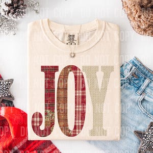 May include: Cream-colored t-shirt with the word "JOY" in a patchwork design. The "J" is red plaid, the "O" is a mix of red and brown plaid, and the "Y" is a beige pattern. The shirt has a crew neck and short sleeves.