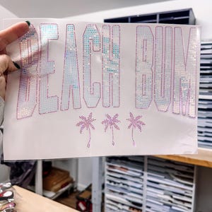 Pode incluir: Uma folha de tecido branco com as palavras "BEACH BUM" escritas em letras grandes, brilhantes, rosa e iridescentes. Três pequenas palmeiras estão abaixo do texto. A folha é segurada por uma mão.