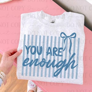 May include: A light grey sweatshirt featuring the words "YOU ARE enough" in blue script. The design includes vertical blue stripes and a blue bow. The sweatshirt is displayed on a pink surface.