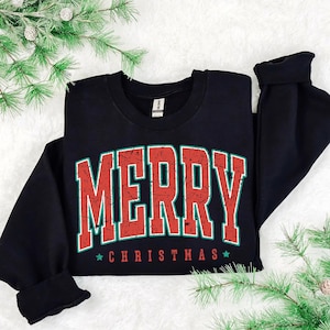Puede incluir: Una sudadera negra con la palabra "MERRY" en letras rojas grandes y desgastadas con contorno verde. Debajo, el texto "CHRISTMAS" está en rojo con estrellas verdes. La sudadera está doblada sobre una superficie blanca con ramas de pino verdes.