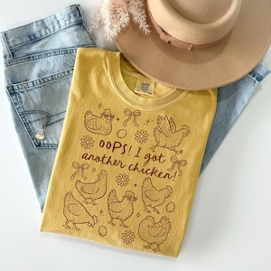 May include: Mustard yellow t-shirt with a brown graphic of chickens, bows, and flowers. The text "OOPS! I got another chicken!" is printed on the front. The shirt is laid on top of light blue jeans and next to a beige hat.