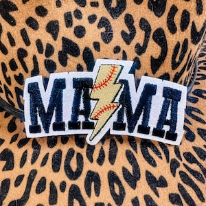 May include: A leopard print hat with a patch that reads "MAMA" in navy blue letters with white trim. A baseball-themed lightning bolt is in the center of the word "MAMA". The hat is brown and black.