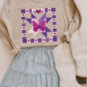 Puede incluir: Sudadera beige con un diseño de mariposa en un patrón de edredón con detalles en morado y rosa. La mariposa está centrada en un diseño de estrella. Combinada con una falda floral azul claro y botas vaqueras blancas.