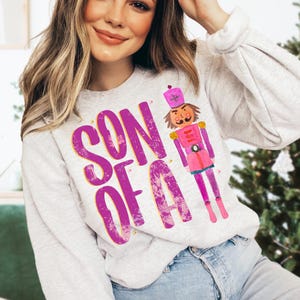 May include: A gray sweatshirt with a pink and yellow graphic design of a nutcracker and the text "SON OF A".