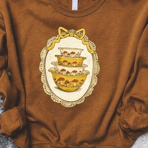 May include: A brown sweatshirt featuring a whimsical design of stacked yellow mixing bowls adorned with red and white mushrooms. The design is framed by a decorative oval with a yellow bow at the top. The sweatshirt is a warm, earthy tone.