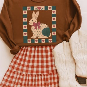 Puede incluir: Una sudadera marrón con un diseño de conejito, lazo rosa y estampado de cuadros. El conejito está enmarcado por un borde verde y beige con detalles de corazón. Combinada con una falda a cuadros roja y blanca y botas vaqueras blancas.