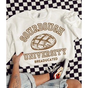Può includere: Felpa bianca con testo marrone che recita "Sourdough University" e "BREADUCATED". Sopra il testo c'è un'illustrazione marrone di una pagnotta di pane a lievitazione naturale.