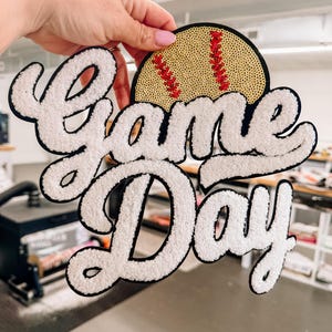 May include: A decorative patch featuring the text Game Day in white chenille letters with a black border. Above the text is a semi-circular gold sequin softball design with red stitching details. The patch has a black felt backing and edge.