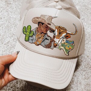 Puede incluir: Gorra de camionero beige con un diseño de vaquero, longhorn y cactus. El diseño utiliza tonos marrones, verdes y beige. La gorra tiene una parte trasera de malla y una visera curva.