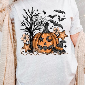 May include: A light gray t-shirt featuring a Halloween-themed graphic. The design includes an orange jack-o'-lantern with a crescent moon and star, bats, stars, flowers, and a witch's hat. The word "Halloween" is written in a script font.