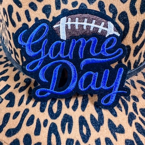 May include: A tan and black leopard print hat with a blue and black patch that says "Game Day" with a football graphic. The hat has a black band around the base.