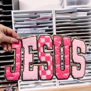Może przedstawiać: Różowa i złota naszywka z napisem „JESUS”. Litery są obrysowane złotym brokatem i mają mieszankę faktur, w tym wzór w szachownicę i detale kokardek. Naszywka jest trzymana przed białą jednostką magazynową.