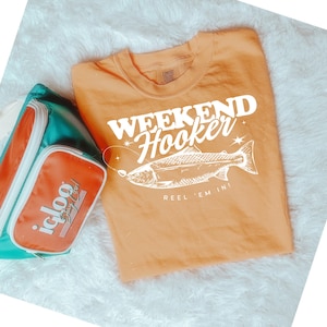 May include: Orange t-shirt with white text that reads "Weekend Hooker Reel 'Em In" and a white illustration of a fish.
