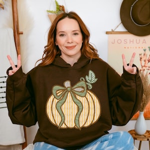 Peut inclure: Un sweat à capuche marron avec un motif de citrouille orné d'un nœud et d'une feuille verts. La citrouille présente des rayures verticales jaunes et blanches. Le sweat est porté par une personne.