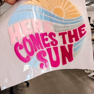 Könnte beinhalten: Ein farbenfroher, kreisförmiger Aufnäher mit dem Text "HERE COMES THE SUN". Das Design zeigt eine gelbe Sonne, blaue und rosa Wellen und den Text in leuchtendem Pink. Der Aufnäher wird in einer Hand gehalten.
