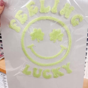 Può includere: Un foglio di plastica trasparente con un design verde neon sfocato. Il design presenta una faccina sorridente con trifogli al posto degli occhi e le parole "FEELIN' LUCKY" in un carattere sfocato abbinato. Il design è probabilmente per un progetto artigianale.