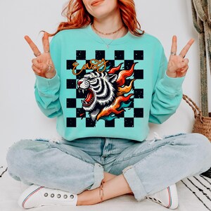 May include: A turquoise blue sweatshirt with a black and white checkered pattern and a tiger head graphic with flames. The graphic also includes the word "easy" in yellow lettering.