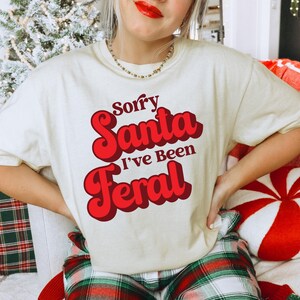 Könnte beinhalten: Cremefarbenes T-Shirt mit dem Text "Sorry Santa I've Been Feral" in roter Retro-Schrift. Das Shirt ist Teil einer weihnachtlichen Bekleidungskollektion. Das Model trägt eine goldene Halskette und roten Lippenstift.