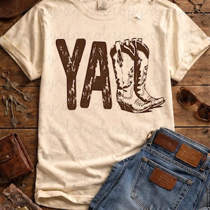 May include: Cream-colored t-shirt with the text "Y'all" and a brown cowboy boot graphic. The shirt is paired with blue jeans, a brown leather wallet, and a cowboy hat, suggesting a Western theme.