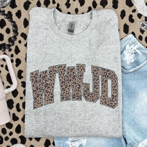 May include: A light gray t-shirt with the letters "WWJD" in a leopard print design. The shirt is folded and placed on a leopard print surface, with denim shorts visible in the background.