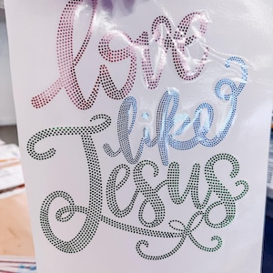 May include: A rhinestone transfer sheet featuring the text Love like Jesus in a cursive script. The word Love is in a pink gradient, like is in a blue gradient, and Jesus is in a green gradient. The design is composed of small, circular rhinestones.