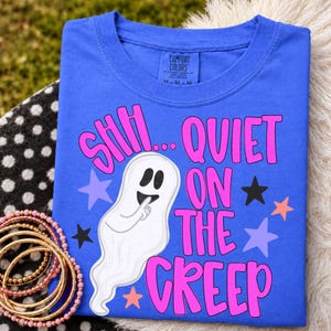 May include: A bright blue t-shirt featuring a white ghost graphic with a finger to its mouth. The text reads SHH... QUIET ON THE CREEP in bold pink letters. The design includes scattered black, light purple, and peach-colored stars across the fabric.