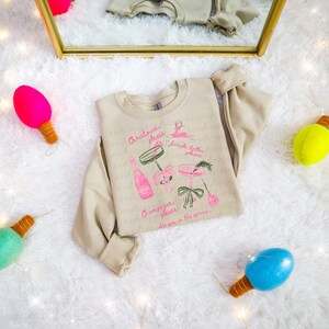 Puede incluir: Sudadera beige con un gráfico rosa y verde de botellas de champán y copas de cóctel. El texto dice "Christmas, please", "Circle bells, please", "Champagne, please" y "See you in the spring..."