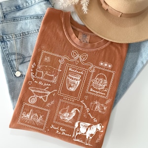 Peut inclure: T-shirt rouille avec un motif sur le thème de la ferme. Le design comprend des illustrations d'un cochon, d'un pot de miel, d'un panier, d'une brouette, d'une poule, d'une vache et le texte "Life is better on the farm" et "Fresh Eggs, No Drama".