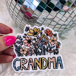 May include: A white embroidered patch featuring a colorful floral arrangement of yellow, orange, and pink flowers above the word GRANDMA, which is stitched in a multi-colored gradient font with blue, yellow, orange, and green letters.
