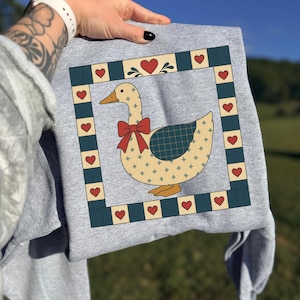 Könnte beinhalten: Graues Sweatshirt mit einem skurrilen Gänsemotiv. Die Gans hat eine rote Schleife und einen karierten Flügel, umrahmt von einem quadratischen Rahmen mit Herzen. Das Design ist im Vintage- und Landhausstil gehalten.