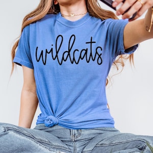 May include: A light blue t-shirt with the word "wildcats" in black script. The shirt is tied in a knot at the waist. The model is wearing blue jeans.