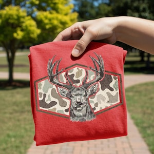 May include: Red t-shirt featuring a detailed illustration of a deer head with large antlers. The deer is set against a camouflage pattern within a hexagonal frame. The shirt is held up by a hand, showcasing the design.